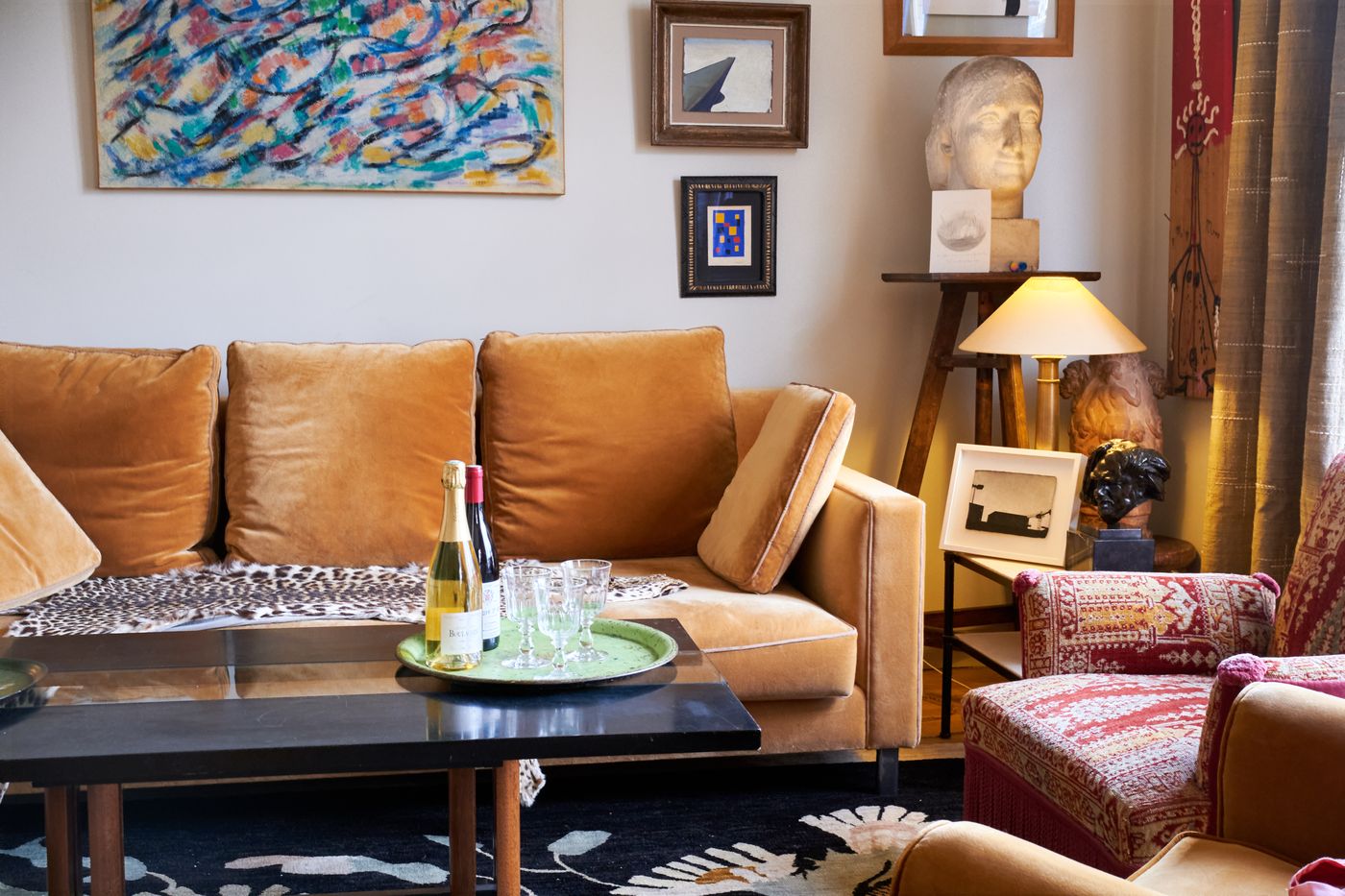 A tray with a bottle and stemmed glasses on a black coffee table in front of an ochre velvet sofa, with paintings and a carved stone head behind
