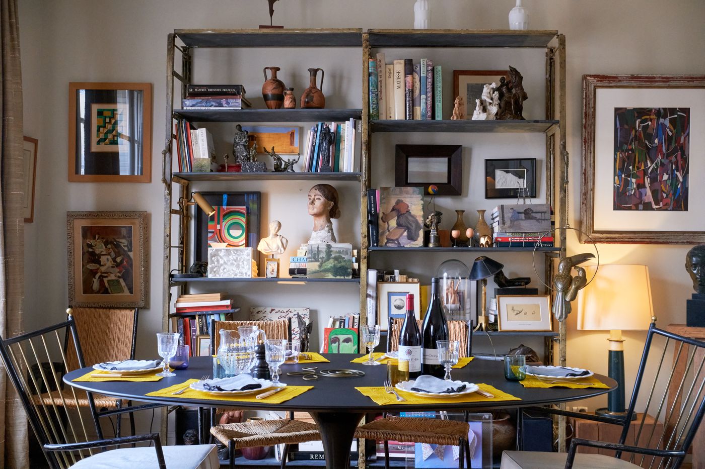 The dining area: a round black table set for dinner in front of tall metal shelves filled with books, ceramics and sculptures, framed works on the walls
