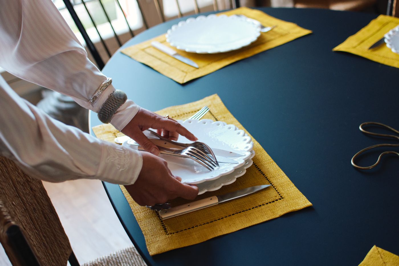 Hands laying scalloped white plates and cutlery on a yellow linen placemat
