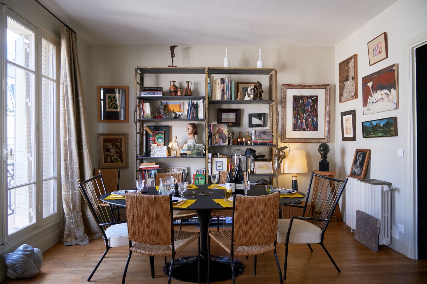 A wide view of the apartment with tall windows, a set dining table and shelves of books and objects