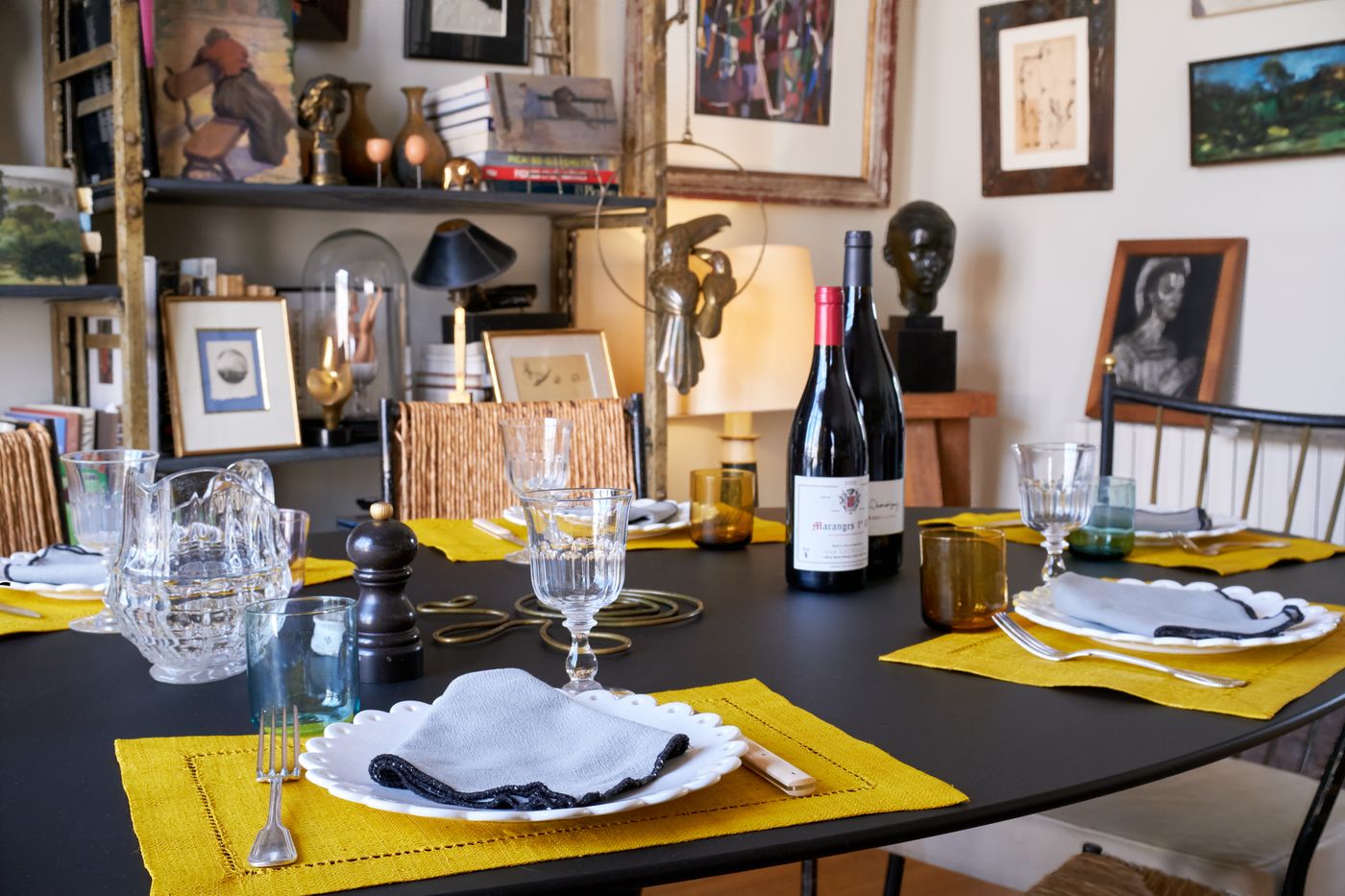 A set dining table with cut-glass glasses, a carafe and two bottles of red wine, framed artworks and a bronze bust behind