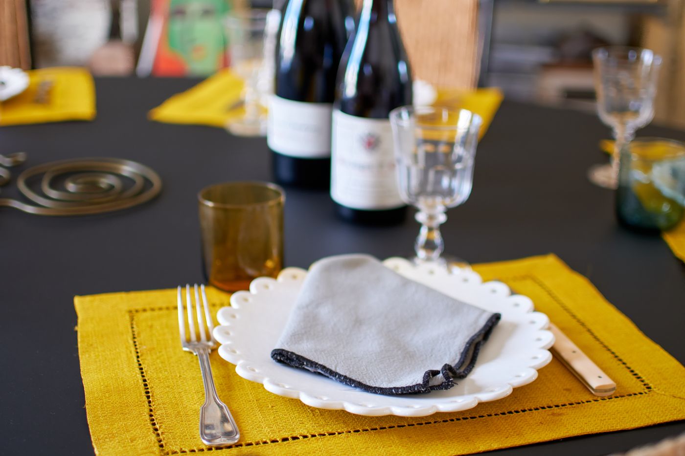 A place setting with a scalloped white plate, a grey linen napkin and a yellow linen placemat, with wine glasses and two bottles of red wine behind